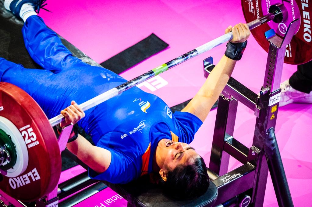 Female para powerlifter in a blue national uniform lifting on the competition bench