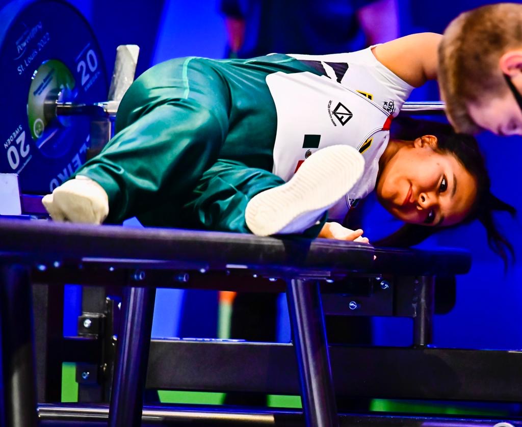 Female para powerlifter wearing a green and white competition uniform moving onto the bench